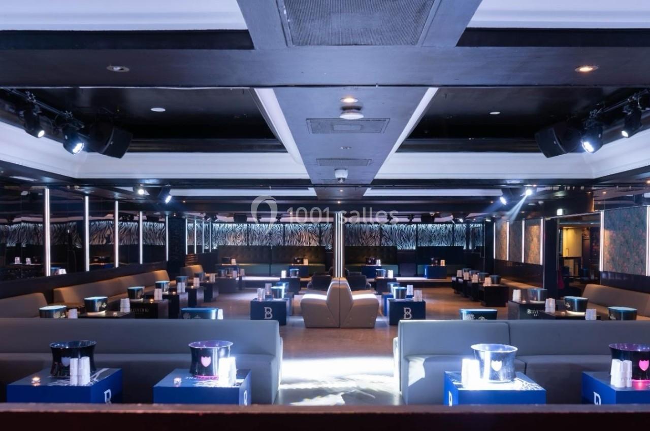 Salle de club avec banquettes grises, tables basses éclairées et ambiance lumineuse tamisée.
