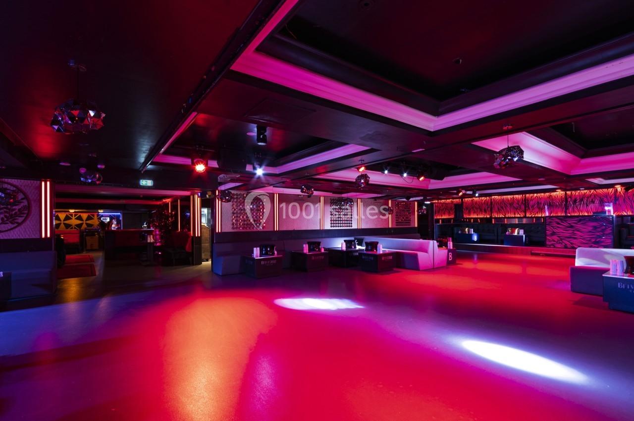 Salle de discothèque vide avec éclairage coloré rouge et violet, banquettes et tables disposées le long des murs.