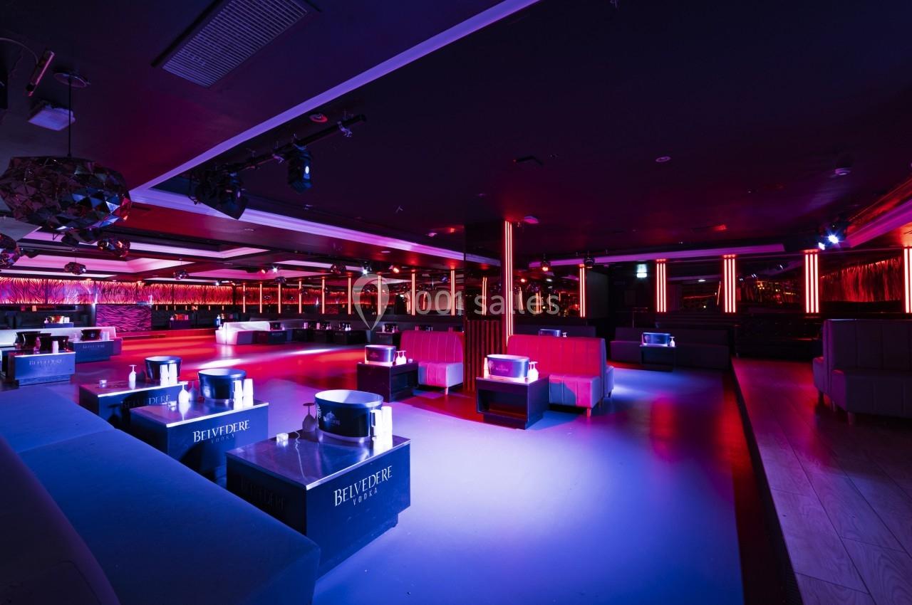 Salle de discothèque avec éclairage tamisé, banquettes rouges, tables basses et piste de danse centrale.