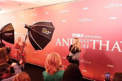 Une femme pose sur un tapis rouge devant un mur promotionnel rose, entourée de photographes et de projecteurs.