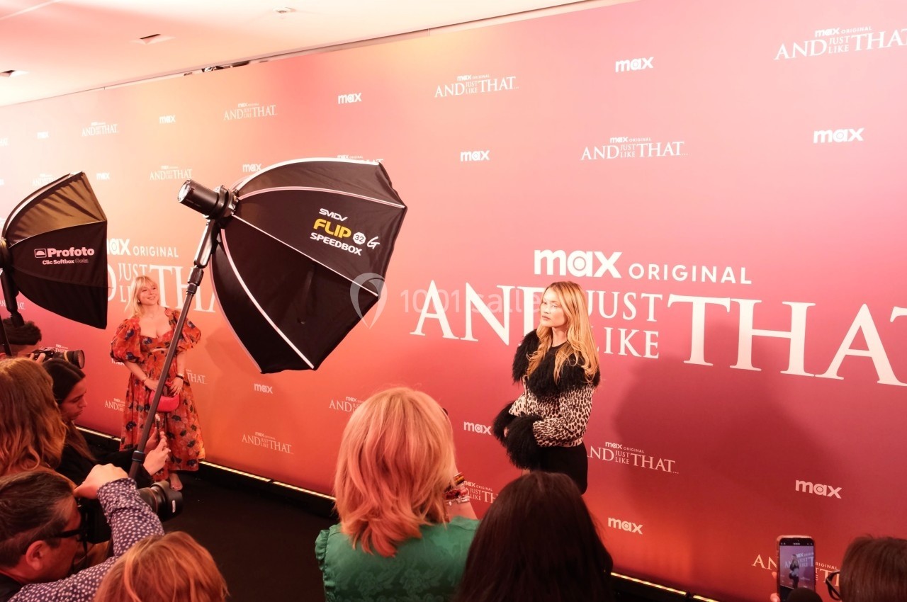 Une femme pose sur un tapis rouge devant un mur promotionnel rose, entourée de photographes et de projecteurs.