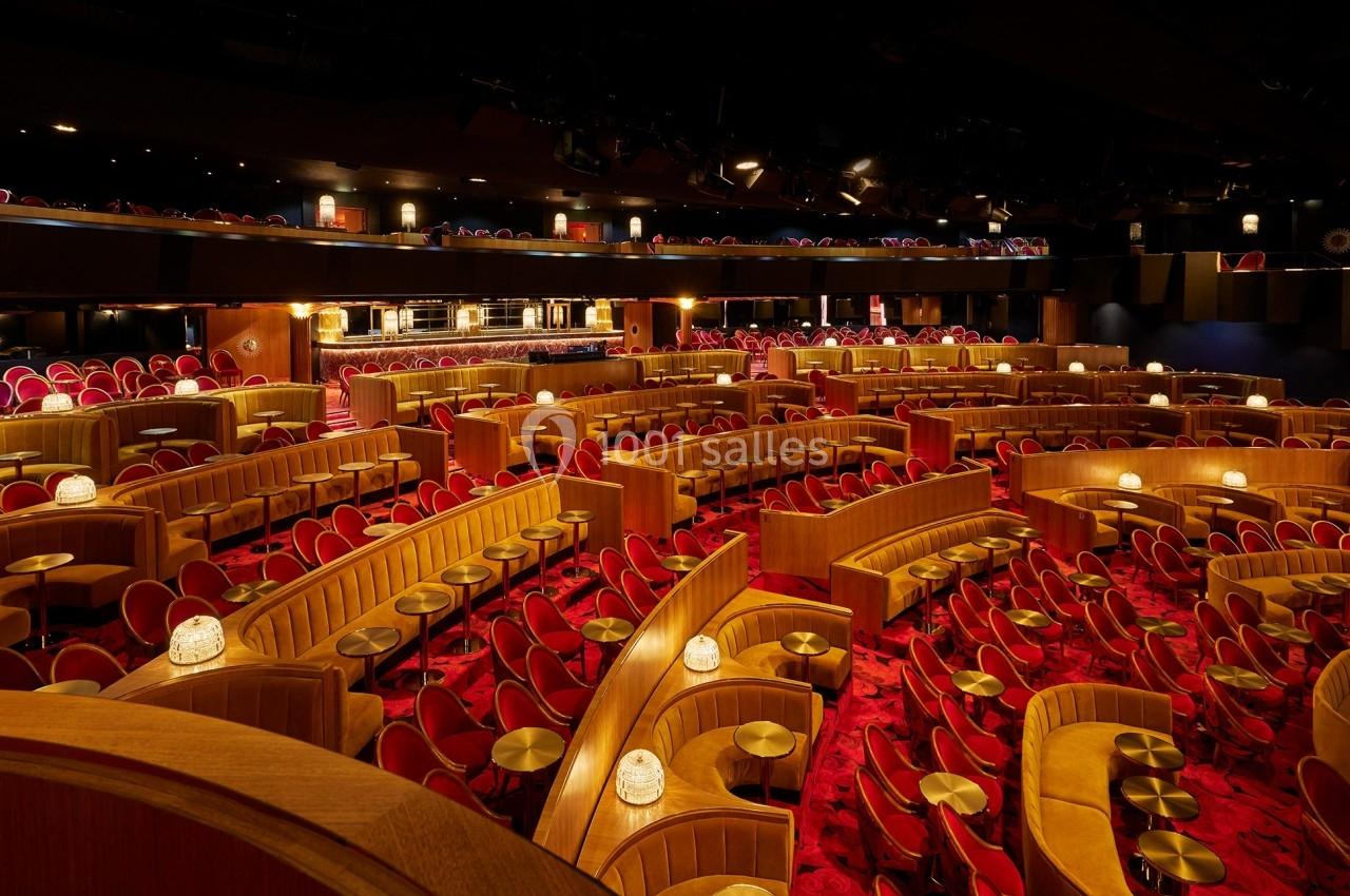Salle de spectacle avec sièges rouges, tables rondes et éclairage tamisé, disposée en gradins circulaires.