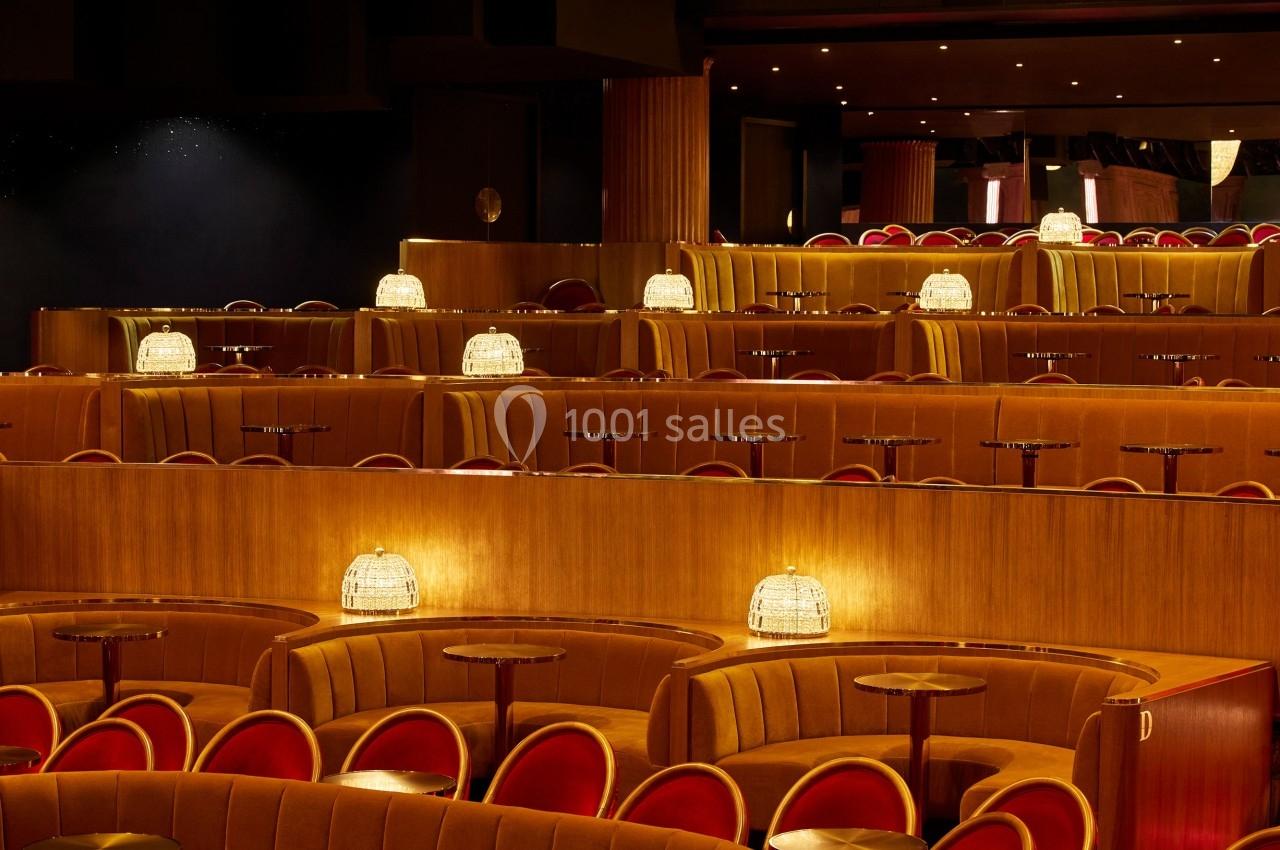 Salle de spectacle avec rangées de banquettes en velours orange, tables rondes et lampes individuelles allumées.