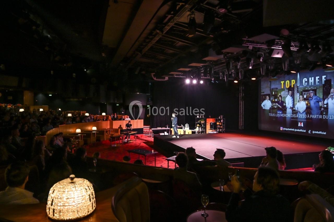 Une scène éclairée avec un orateur devant un public dans une salle de spectacle, écran projetant ’Top Chef’.