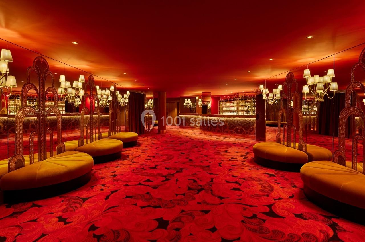 Salle au décor rouge vif avec moquette ornée de motifs, banquettes arrondies et miroirs reflétant des lustres.