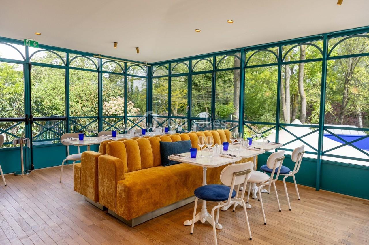 Salle lumineuse avec baies vitrées, mobilier mêlant chaises blanches et banquettes jaunes, vue sur un jardin verdoyant.