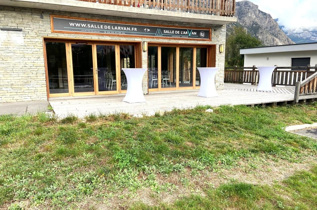 Terrasse en bois avec tables hautes drapées de blanc devant une salle vitrée, entourée de montagnes.