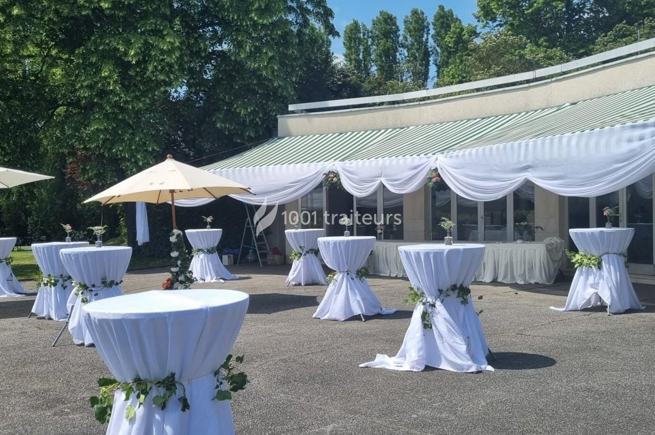 Tables hautes drapées de blanc décorées de feuillage, disposées en extérieur devant un bâtiment avec auvent rayé.