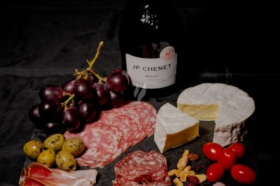 Plateau de charcuterie et fromages avec fruits, légumes et une bouteille de vin rouge, présenté sur une table en extérieur.