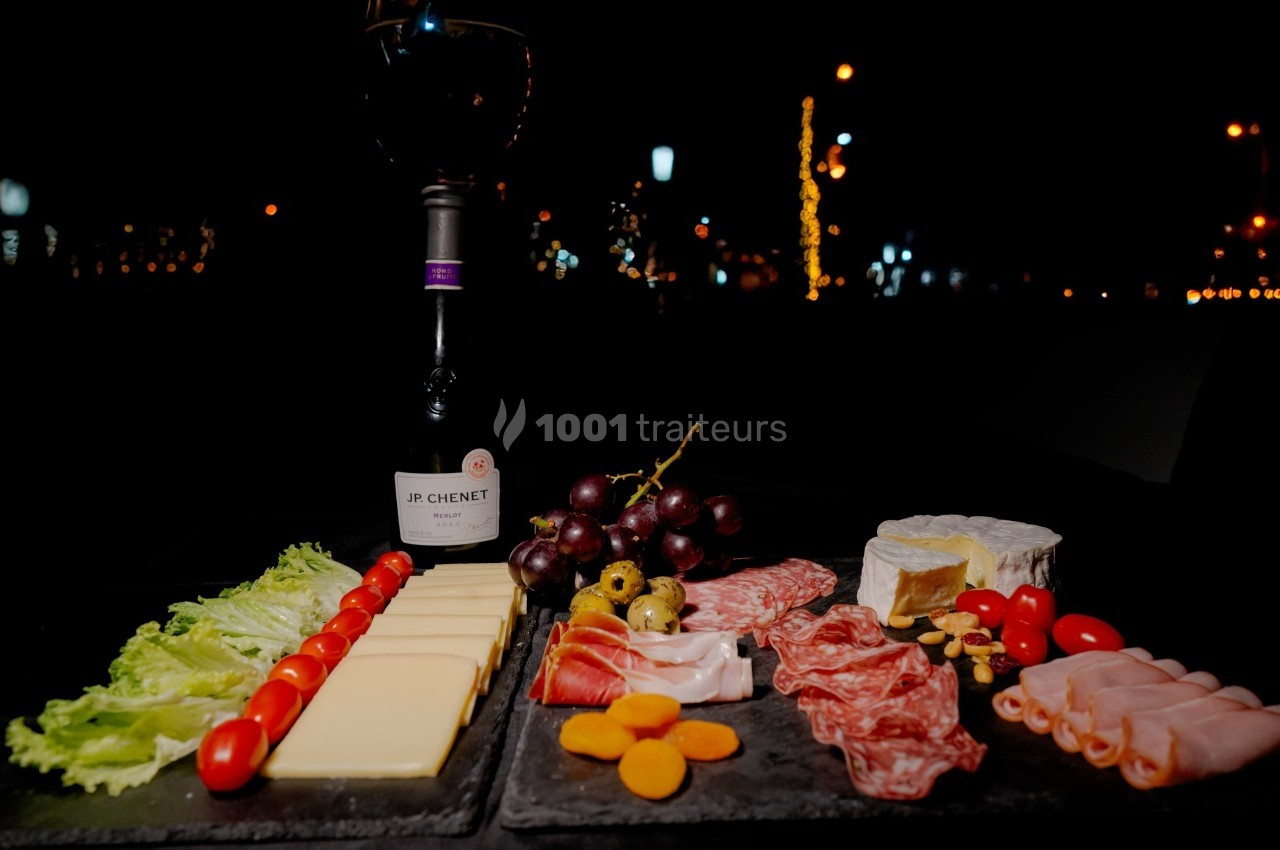 Plateau de charcuterie et fromages avec fruits, légumes et une bouteille de vin rouge, présenté sur une table en extérieur.