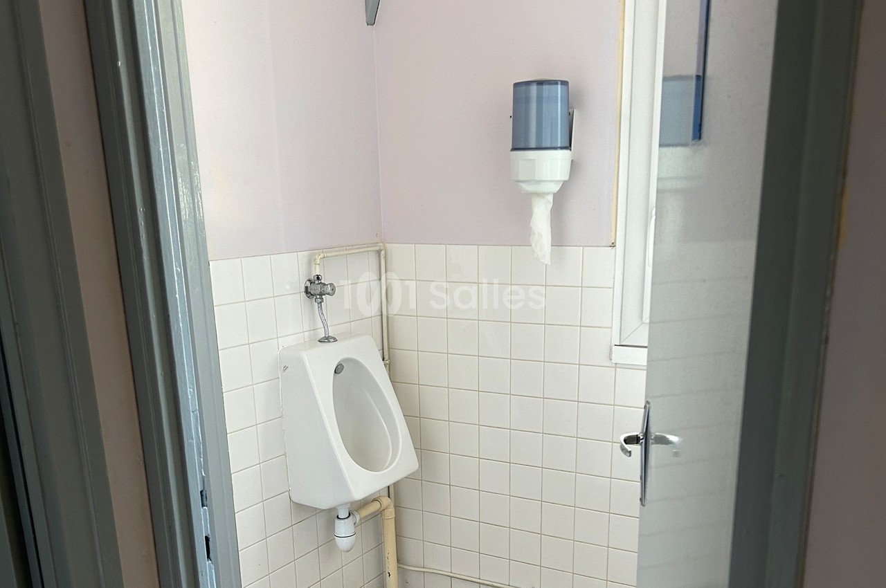 Urinoir mural blanc dans une petite pièce carrelée avec un distributeur de papier fixé au mur.