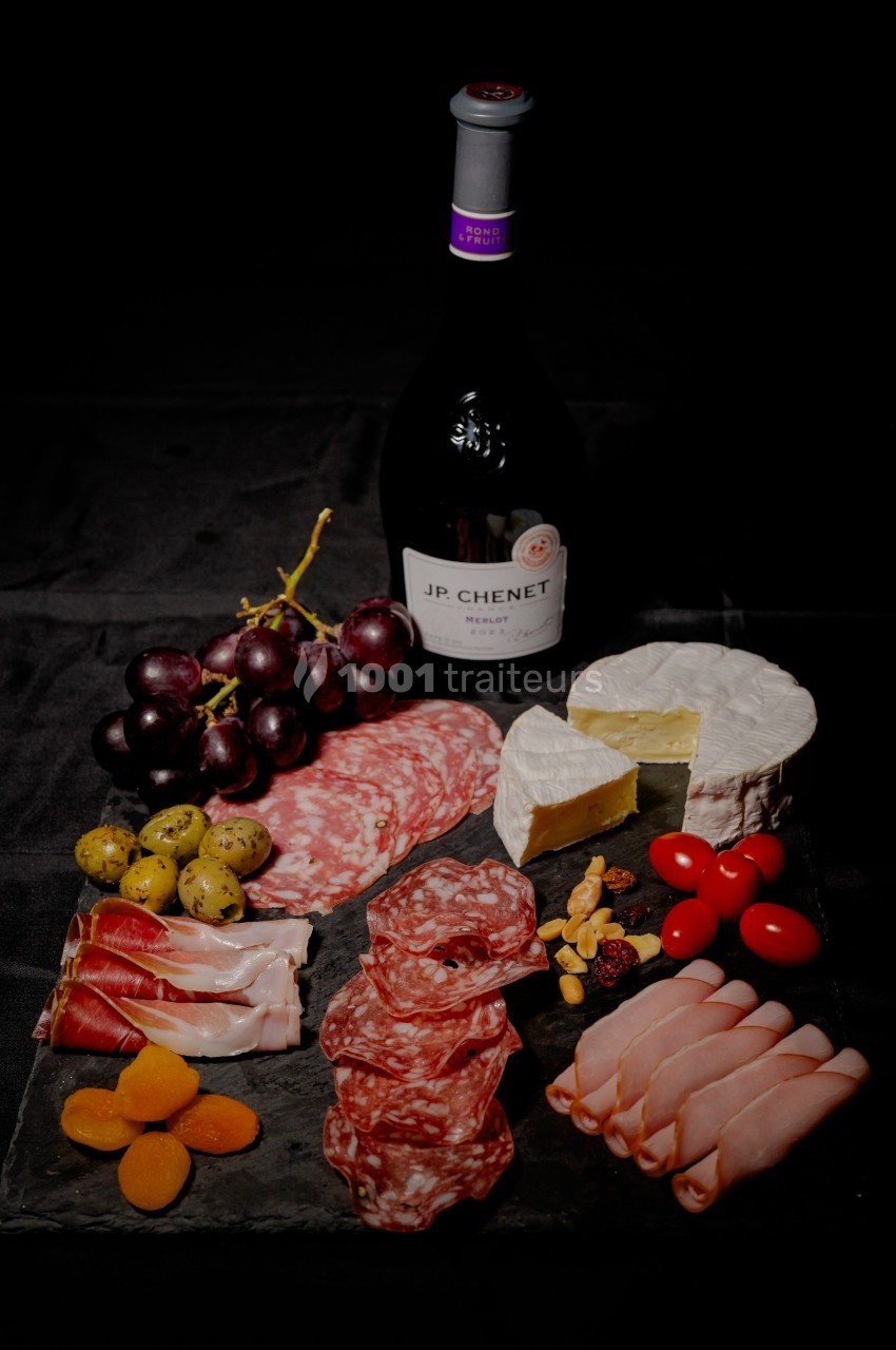 Plateau de charcuterie avec fromages, fruits, olives, noix et une bouteille de vin rouge sur fond noir.