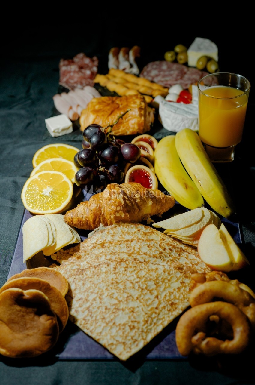 Assortiment de viennoiseries, fruits, charcuteries, fromage et jus d'orange sur une table sombre.
