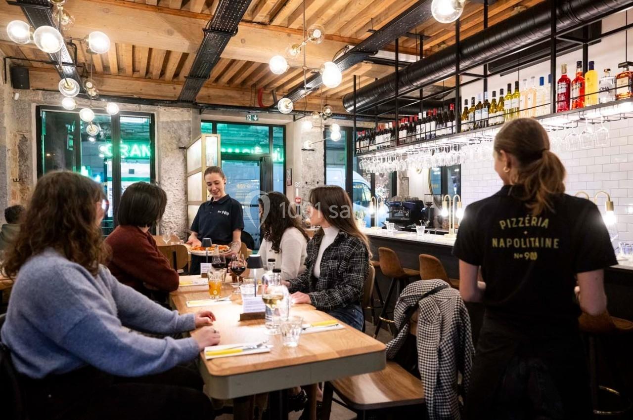 Des clients discutent autour de tables en bois dans une pizzeria animée avec un décor moderne et chaleureux.