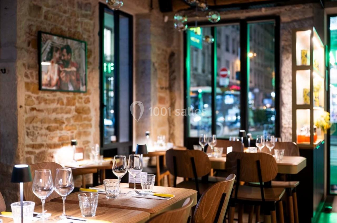 Salle de restaurant chaleureuse avec murs en pierre, tables dressées et vue sur la rue éclairée à travers de grandes…