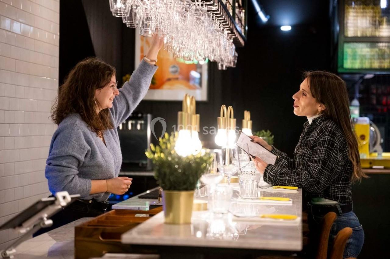 Deux femmes discutent au comptoir d'un bar, l'une tendant la main vers des verres suspendus.