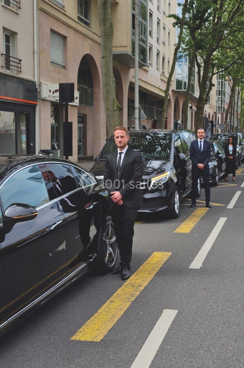 Des chauffeurs en costume debout devant une file de véhicules noirs garés le long d'une rue bordée d'arbres.
