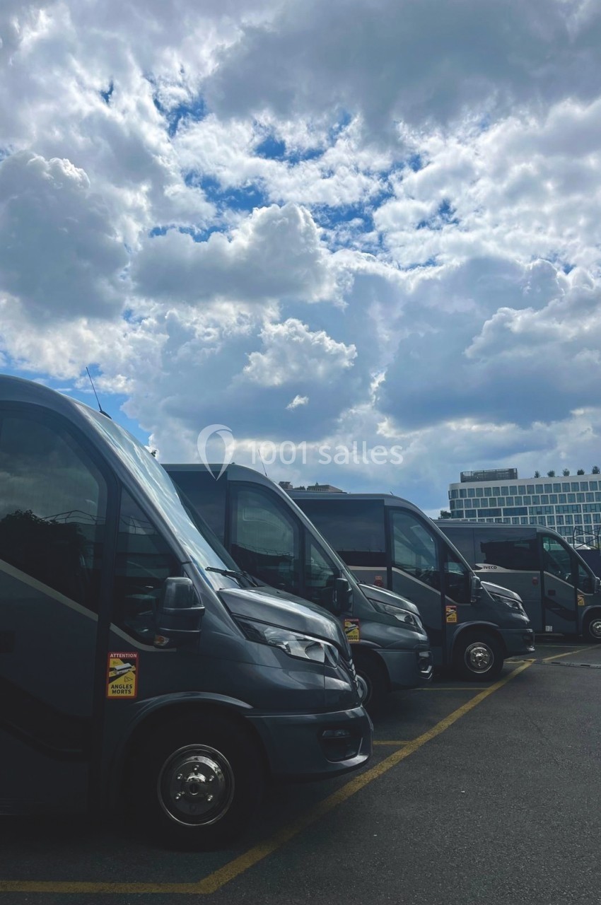 Alignement de minibus gris stationnés sur un parking sous un ciel nuageux.