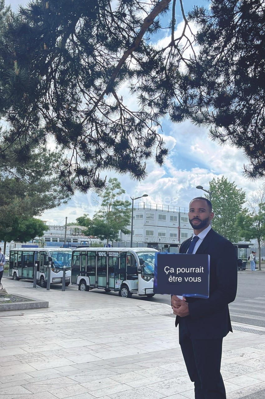 Un homme en costume tient une pancarte ’Ça pourrait être vous’ devant une rangée de bus électriques en extérieur.