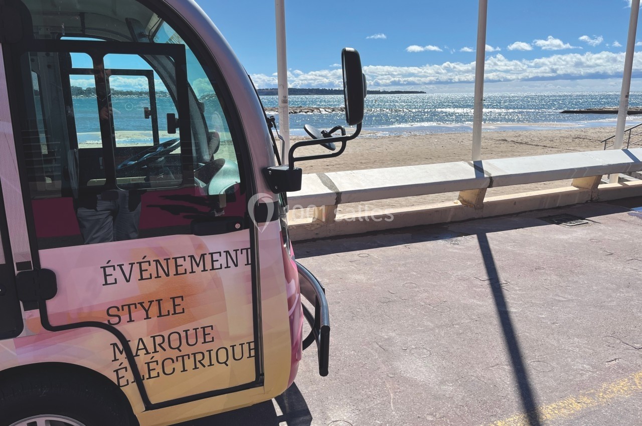 Minibus électrique stationné près d'une plage ensoleillée avec vue sur la mer et un ciel partiellement nuageux.