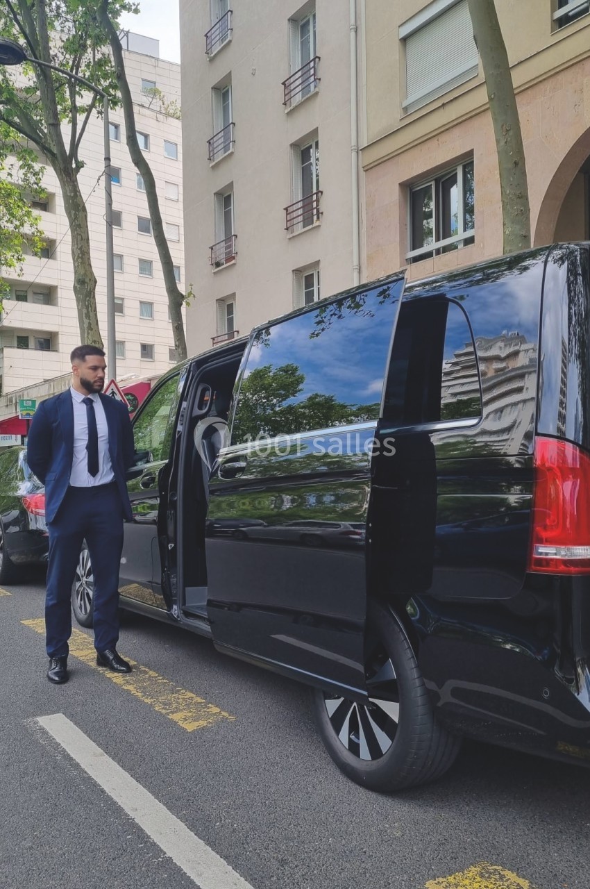 Un homme en costume se tient près d'une voiture noire avec une porte latérale ouverte, stationnée en ville.