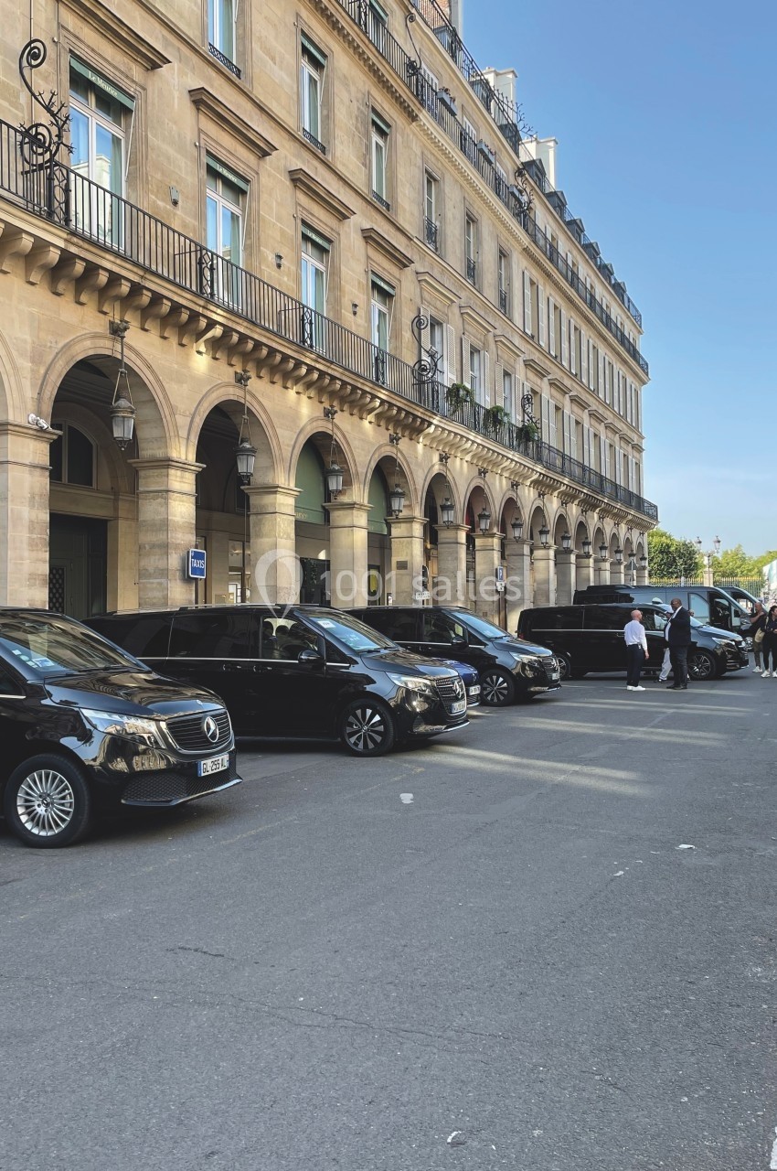 Alignement de véhicules noirs stationnés devant un bâtiment en pierre avec arcades, sous un ciel dégagé.