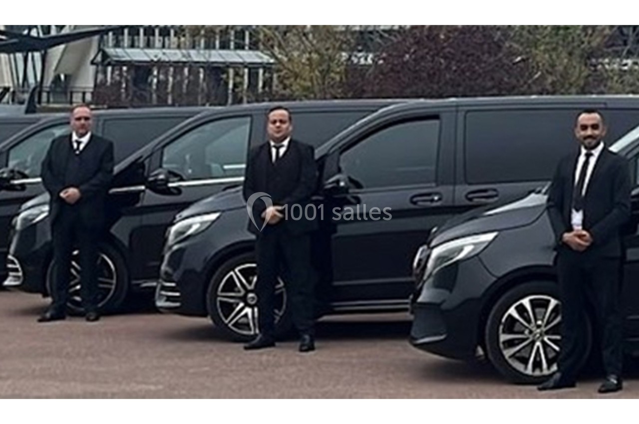 Trois hommes en costume noir se tiennent debout devant une rangée de vans noirs garés.