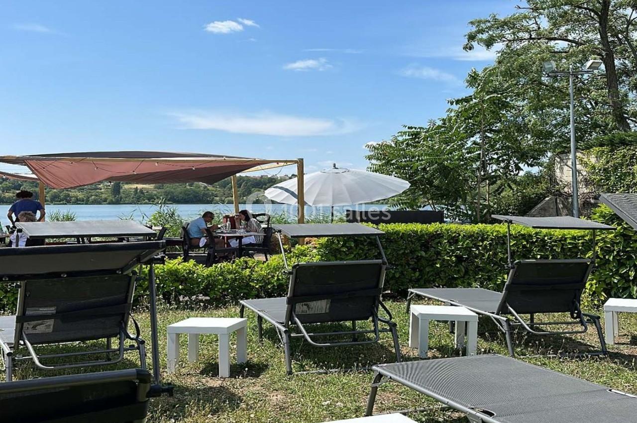 Des chaises longues et des tables installées près d'un lac, avec des parasols et des arbres en arrière-plan.