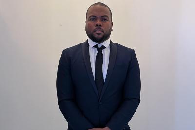 Homme en costume sombre et cravate noire, debout devant un mur blanc.