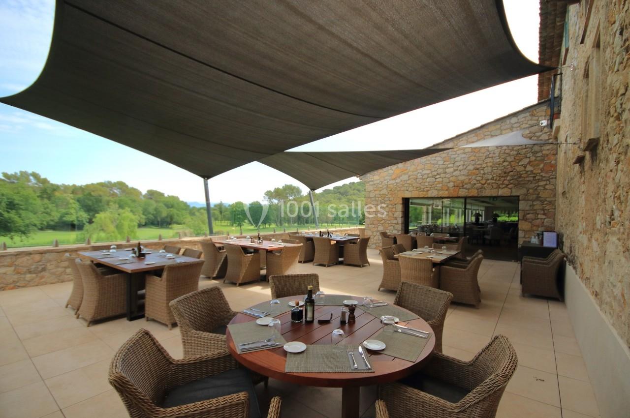 Terrasse extérieure avec tables en bois, chaises en rotin et voiles d'ombrage, donnant sur un paysage verdoyant.