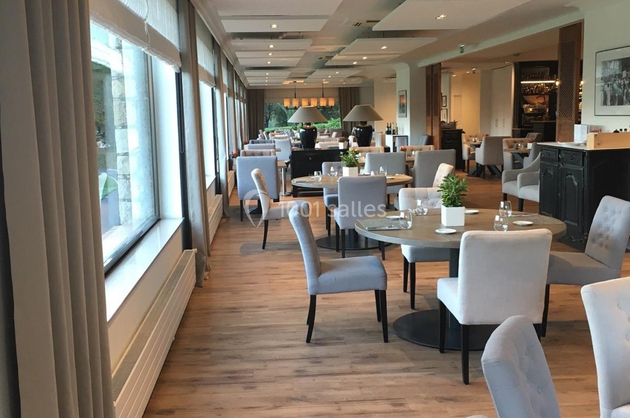 Salle de restaurant lumineuse avec grandes baies vitrées, tables rondes dressées et chaises rembourrées grises.