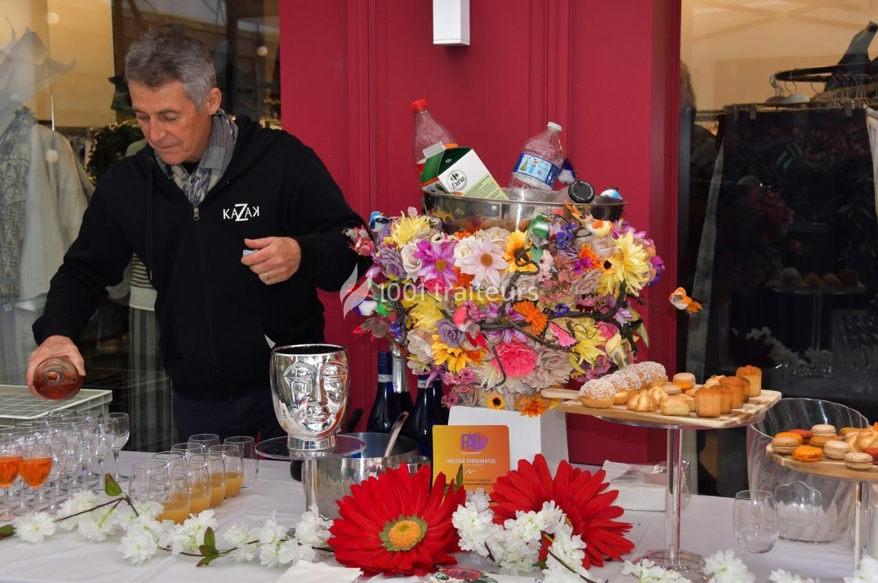 Un homme sert une boisson sur une table décorée de fleurs, avec des desserts et des bouteilles en arrière-plan.