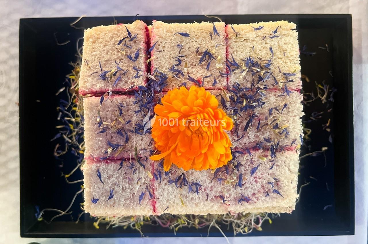 Gâteau carré décoré de fleurs comestibles violettes et orange, présenté sur un plateau noir.