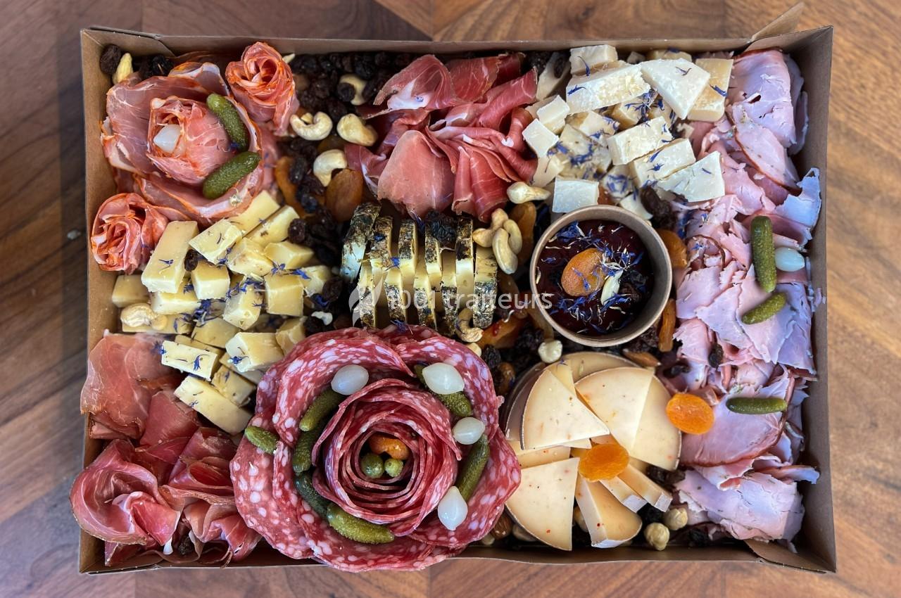 Plateau de charcuterie et fromages variés avec cornichons, fruits secs, noix et confiture dans une boîte en carton.
