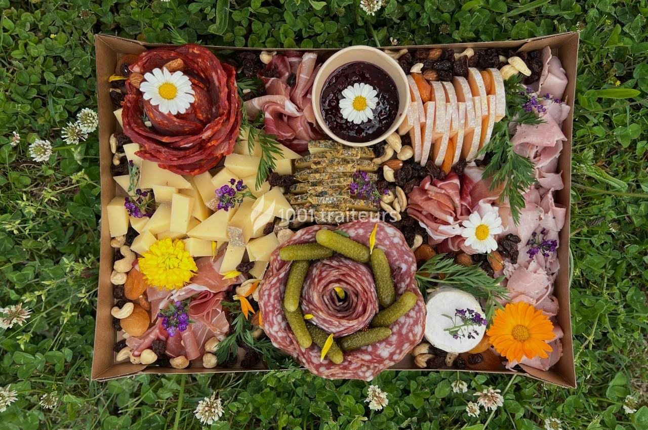 Plateau de charcuterie et fromages variés avec fruits secs, cornichons et décorations florales, posé sur de l'herbe.