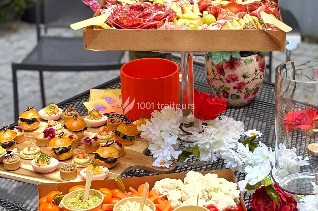 Plateau de charcuterie, mini-burgers et légumes frais disposés sur une table décorée de fleurs.