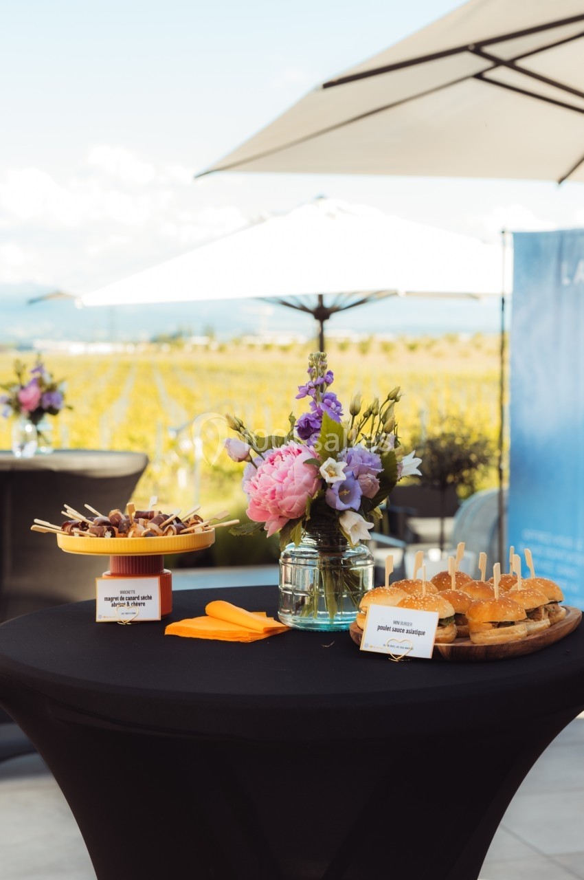 Table haute avec apéritifs, fleurs en vase et parasols en arrière-plan sur une terrasse ensoleillée.