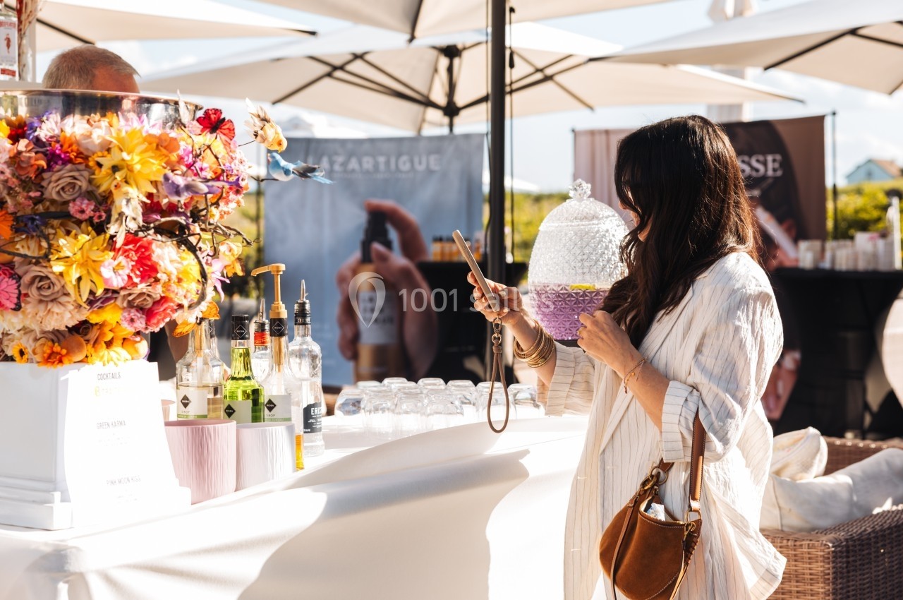 Une femme prend en photo une boisson violette sur un stand extérieur décoré de fleurs et de bouteilles.