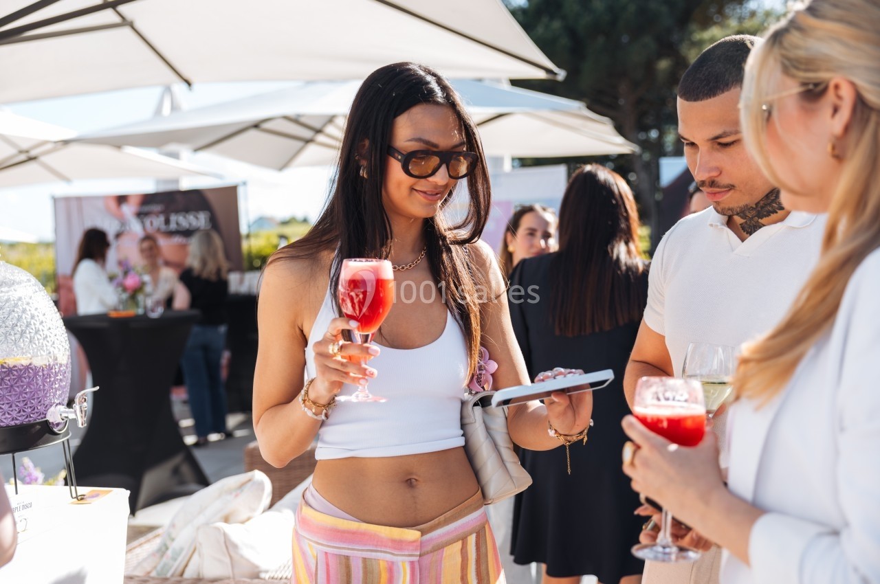 Une femme tenant un verre de cocktail rouge discute avec deux personnes lors d'un événement en extérieur.