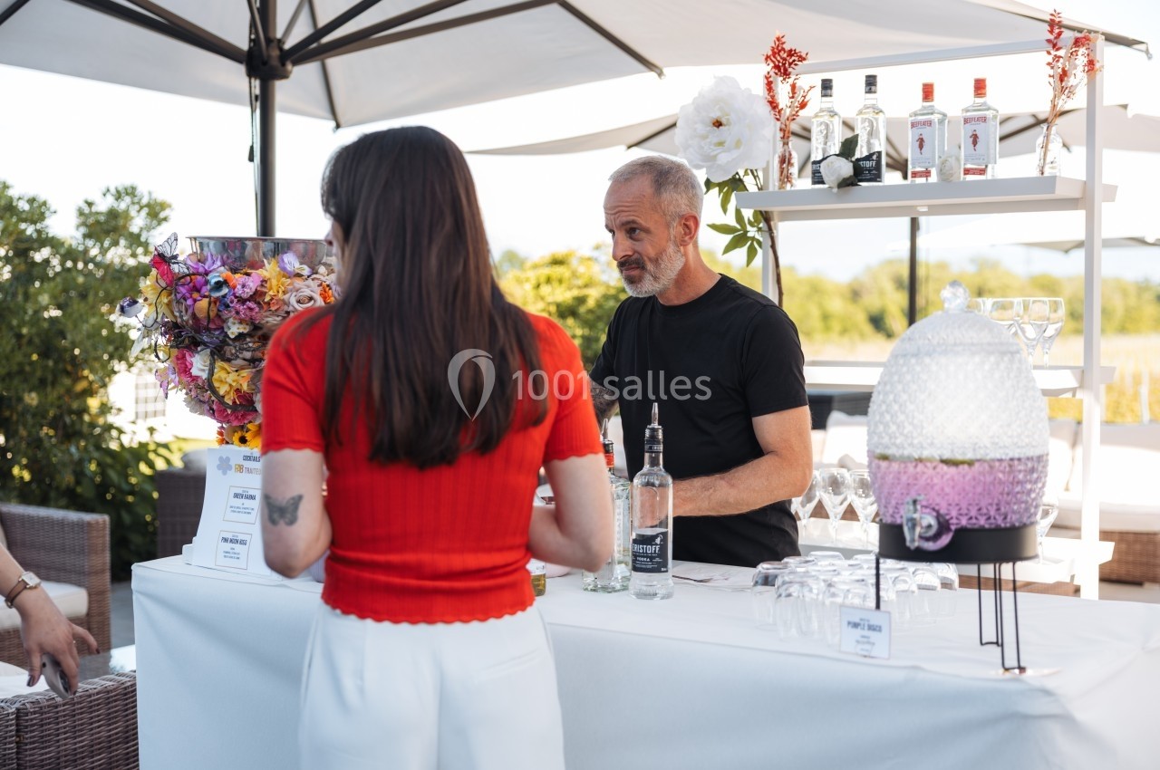 Un barman sert une cliente à un stand en plein air, avec des bouteilles et des verres disposés sur une table.