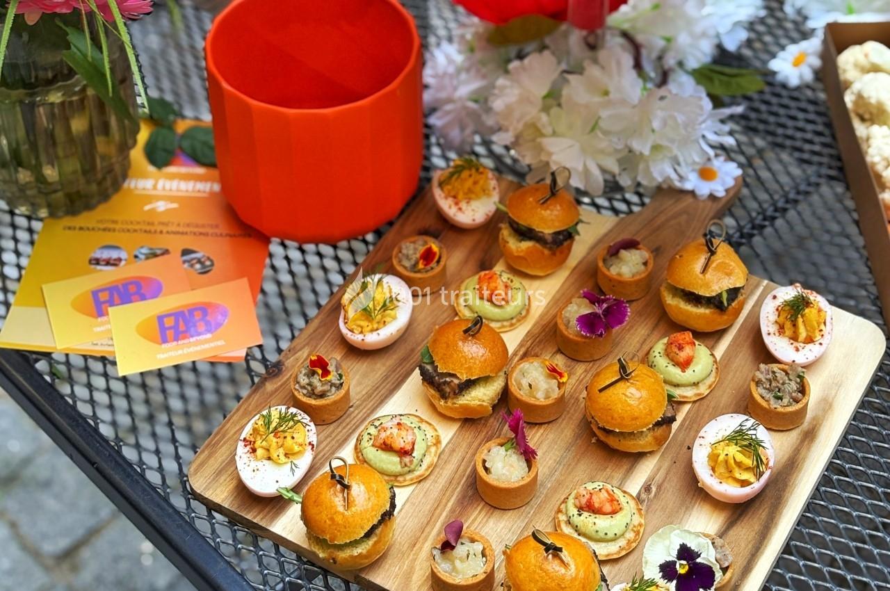 Plateau en bois avec des amuse-bouches variés, incluant mini-burgers, tartelettes et œufs garnis, posé sur une table en…