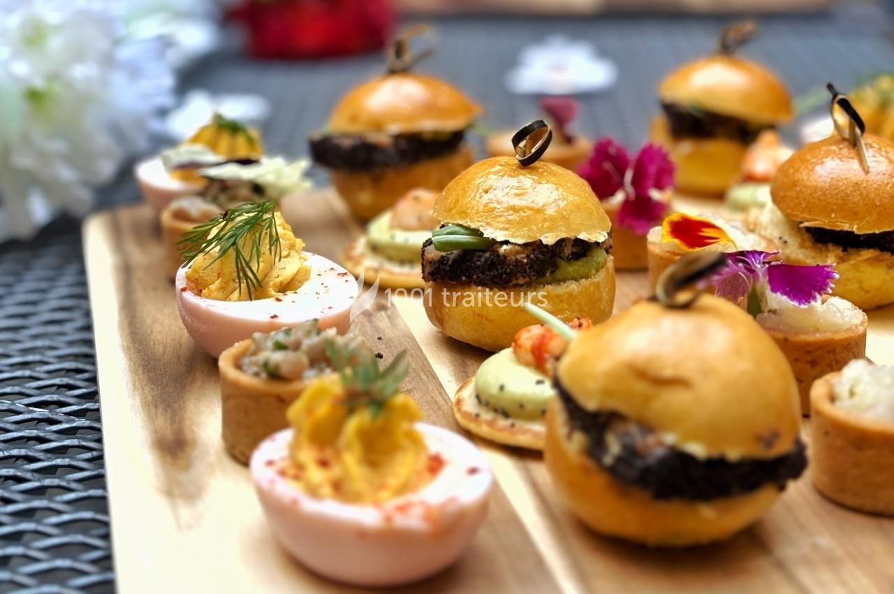 Plateau en bois garni de mini-burgers, bouchées salées et œufs farcis décorés d'herbes et de fleurs comestibles.