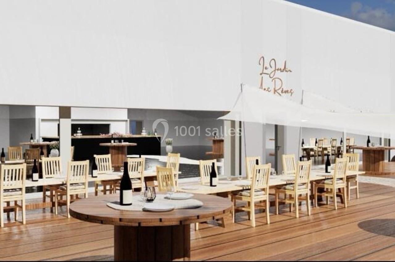 Terrasse en bois avec tables et chaises en bois clair, bar en arrière-plan et voiles d'ombrage blanches.