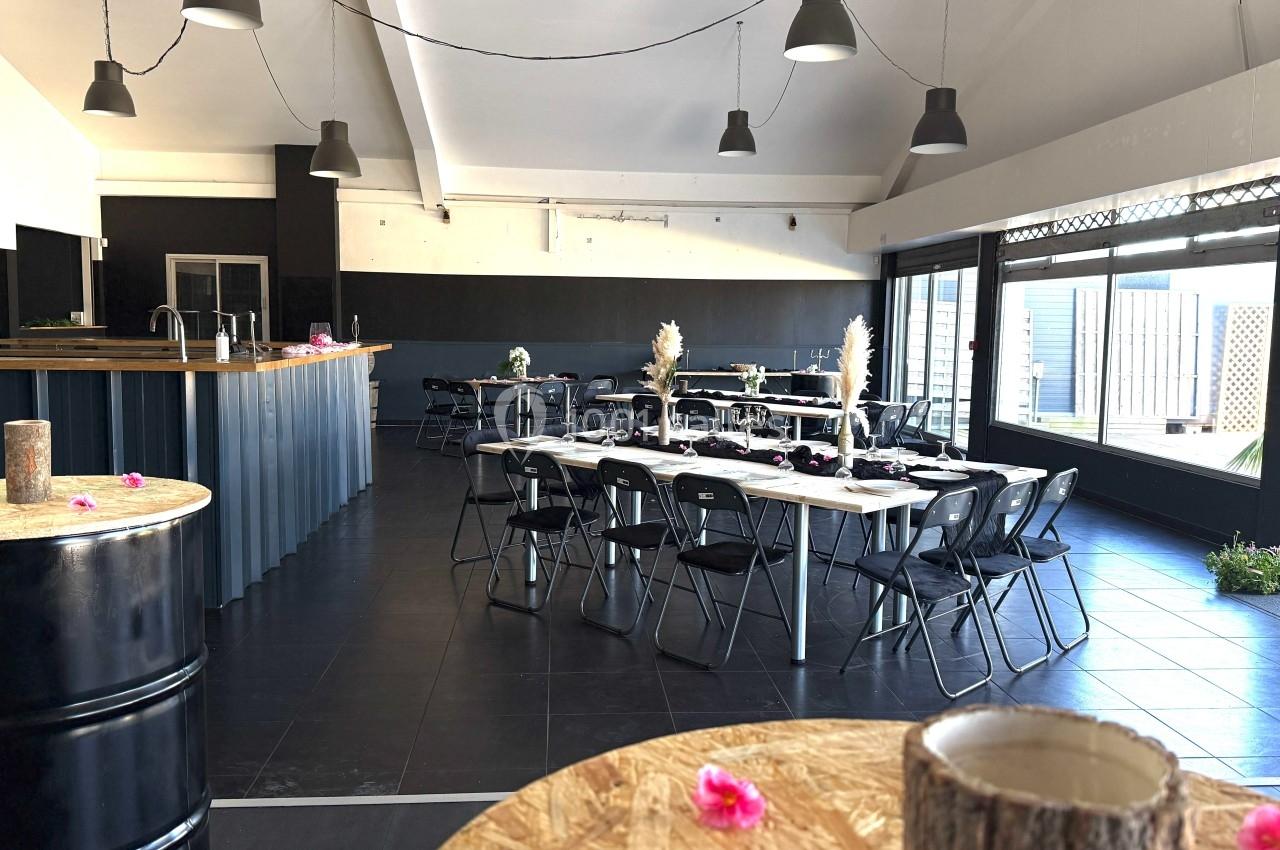 Salle lumineuse aménagée pour un événement, avec tables dressées, chaises noires et décoration sobre.