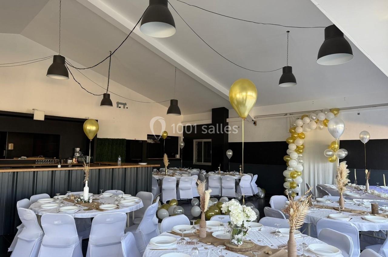 Salle décorée pour un événement avec tables dressées, chaises blanches, ballons dorés et arche de ballons en arrière-plan.
