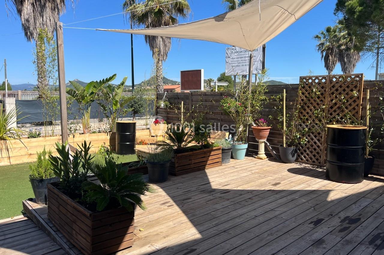 Terrasse en bois avec des plantes en pots, des voiles d'ombrage et des palmiers en arrière-plan sous un ciel bleu.