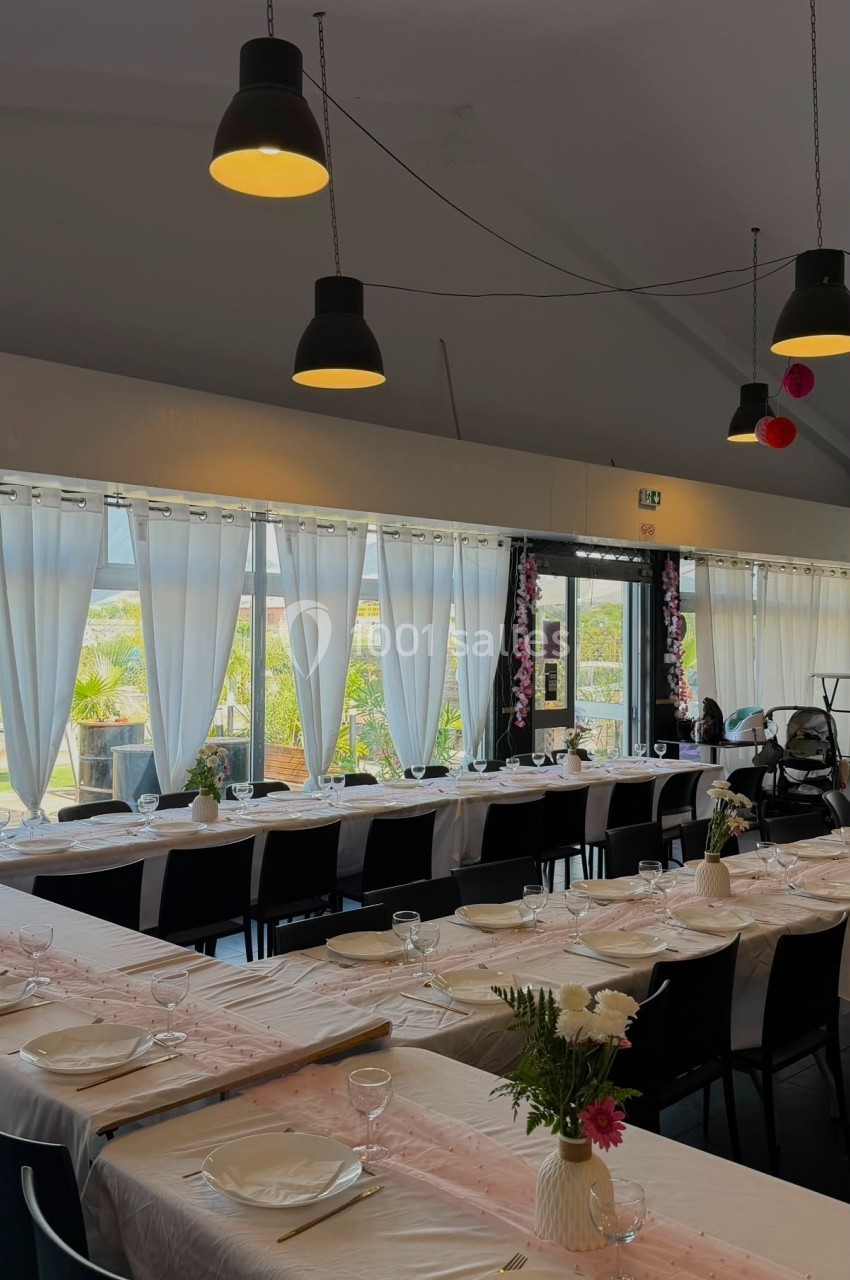 Salle décorée pour un repas, tables alignées avec nappes blanches, vaisselle et fleurs, grandes fenêtres en arrière-plan.