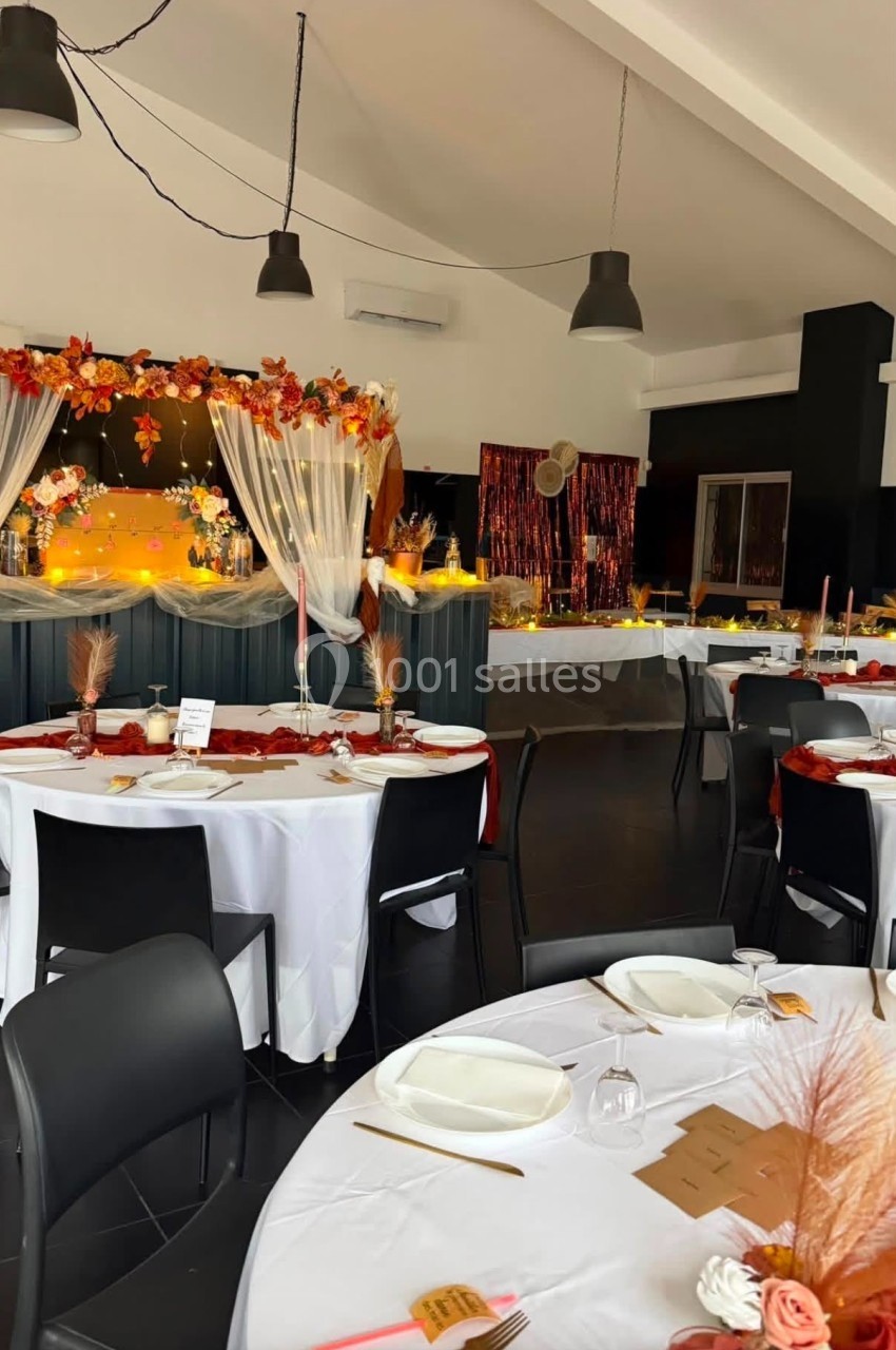 Salle décorée pour un événement avec tables dressées, nappes blanches, décorations automnales et guirlandes lumineuses.