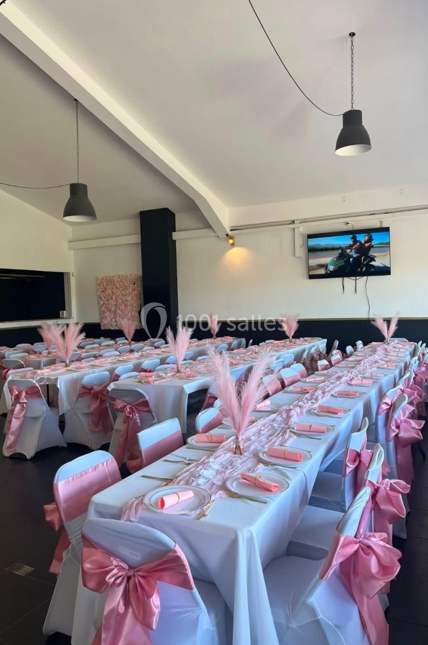 Salle décorée pour un événement, avec tables alignées, nappes blanches, nœuds roses et plumes en centre de table.