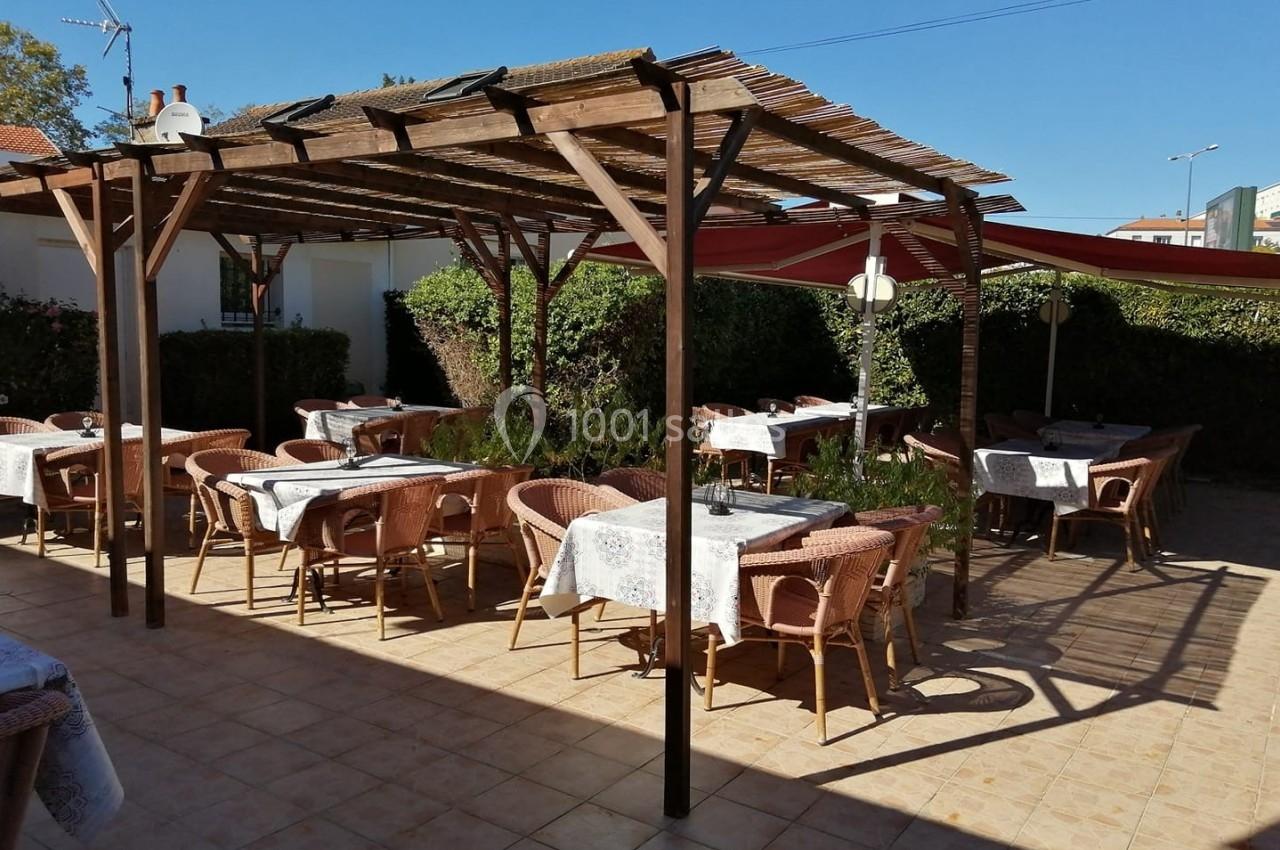 Terrasse extérieure avec tables et chaises en osier sous une pergola en bois, entourée de haies et de parasols.